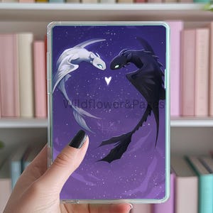 May include: A clear phone case with a purple background and a white and black dragon design. The dragons are facing each other with a heart between them. The text "Wildflower & Pages" is printed on the case.