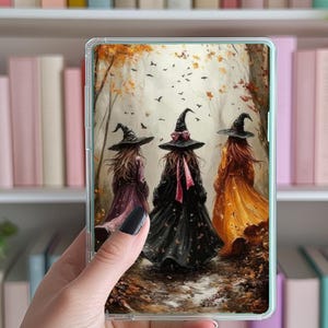 May include: A clear phone case featuring a painting of three witches walking in a forest. The witches wear black hats and colorful dresses in shades of purple, black, and orange. The background shows autumn foliage and flying birds.