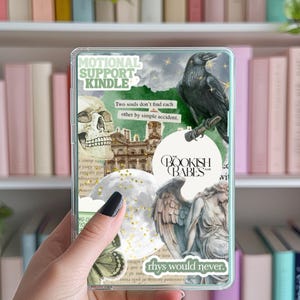 May include: A clear plastic phone case with a collage of images, including a raven, a skull, a building, a cloud with gold glitter, an angel, and a butterfly. The case has a sticker that says "MOTIONAL SUPPORT KINDLE" and another sticker that says "Two souls don't find each other by simple accident." There is also a sticker that says "BOOKISH BABES" and another sticker that says "rhys would never."