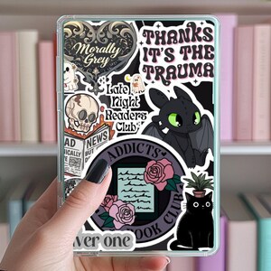 May include: A clear phone case with various stickers, including a black dragon, a skull, a ghost, a book club logo, a cat, and a rose. The text on the stickers includes "Thanks it's the trauma", "Morally Grey", "Late Night Readers Club", "Addicts Book Club", and "Never One".