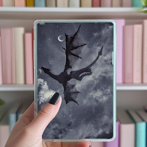 May include: A clear phone case featuring a black and white image of a dragon soaring through a cloudy night sky. The dragon has large wings and a long tail. The sky is dotted with stars and a crescent moon.