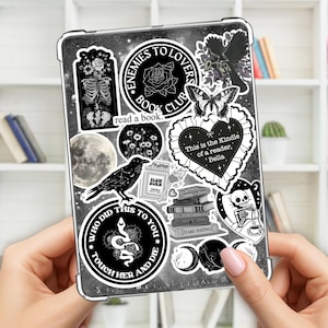 May include: A clear tablet case adorned with various black and white stickers. Designs include a raven, a skeleton, a snake, a moon, and text that reads "Enemies to Lovers Book Club" and "Who Did This To You".