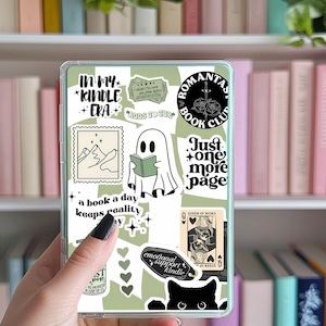 Könnte beinhalten: Eine transparente Hülle für einen E-Reader, verziert mit Aufklebern. Die Aufkleber zeigen Sätze wie "In My Kindle Era", einen Geist, der ein Buch liest, und eine schwarze Katze. Die Hülle wird vor einem Bücherregal mit Büchern gehalten.