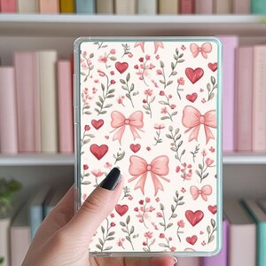 May include: A clear phone case with a pink and white floral design featuring hearts and bows. The design is repeated throughout the case.