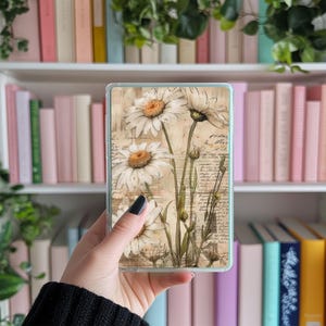 Könnte beinhalten: Eine transparente Handyhülle mit einem Blumendesign mit weißen Gänseblümchen und grünen Stielen auf einem Vintage-Hintergrund mit verblassten Text.