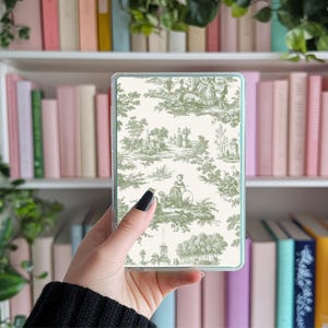 May include: A clear phone case with a green and white toile pattern. The pattern features a repeating design of trees, flowers, and people.