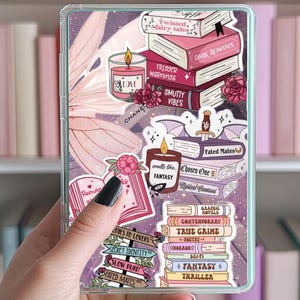 May include: A clear phone case with a pink and purple background. The case is decorated with stickers of books, candles, and other bookish items. The stickers include the words "Fated Mates", "Chosen One", "True Crime", "Fantasy", "Thriller", "Dark Romance", "Trigger Warnings", "Smutty Vibes", "Twisted Fairy Tales", "Smells Like Fantasy", "Romances to Lovers", "Secret Identity", "Slow Burn", "Fated Mates", and "Graphic Novels".