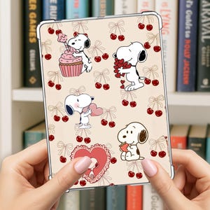 May include: A clear tablet case featuring a repeating pattern of Snoopy characters, cherries, and bows on a beige background. The case is held by a person. The Snoopy illustrations include Snoopy holding a cupcake, hearts, and strawberries.