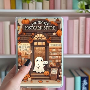 May include: A phone case featuring a whimsical illustration of "Mr. Ghost Postcard Store." The design includes a ghost holding an envelope, pumpkins, and autumn foliage. The store's facade is detailed with a sign and various postcards.