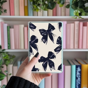 Peut inclure: Une coque de téléphone transparente avec un motif de nœuds bleu marine sur fond crème. Les nœuds sont stylisés avec des détails au pinceau. La coque est tenue devant une bibliothèque remplie de livres.