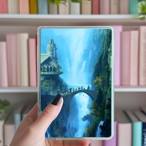 Könnte beinhalten: Eine transparente Handyhülle mit einer Fantasy-Landschaft mit Wasserfall, Brücke und Gebäude auf einer Klippe. Die Hülle wird in einer Hand gehalten, mit einem Bücherregal im Hintergrund.