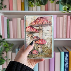 Op de afbeelding: Een doorzichtig telefoonhoesje met een aquarel illustratie van rood-wit gestippelde paddenstoelen. Het ontwerp bevat een vintage papieren achtergrond met handschrift en groen gras. De hoes wordt in een hand gehouden.