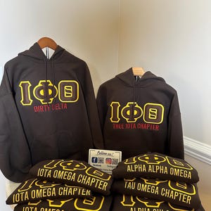 Peut inclure: Des sweats à capuche et des sweat-shirts bruns avec des lettres dorées et rouges. Les sweats à capuche affichent "IΦΘ" et des noms de chapitres comme "Dirty Delta" et "Thee Iota Chapter". Les sweat-shirts sont empilés, montrant "Iota Omega Chapter".