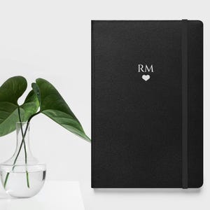 Op de afbeelding: Een zwart lederen gebonden notitieboek met een witte elastische sluiting en de initialen "RM" met een hart boven de letters.