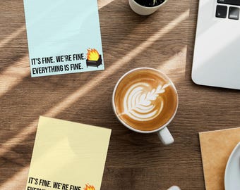 May include: Two sticky notes, one light blue and one yellow, with the text "IT'S FINE. WE'RE FINE. EVERYTHING IS FINE." and a graphic of a burning grill. The image also includes a cup of coffee, a laptop, and a small plant.