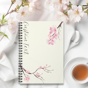 Op de afbeelding: Een wit spiraalnotitieblok met een crèmekleurige omslag met een aquarelontwerp van roze kersenbloesems op takken. De tekst "Custom Text" is in zwarte cursieve letters op de omslag geschreven.
