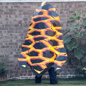Könnte beinhalten: Ein tragbarer Umhang mit einem geometrischen, ineinandergreifenden Sechseckmuster in Schwarz, Orange und Gelb, das an fließende Lava erinnert. Das Kleidungsstück besteht aus mehreren starren, geschichteten Paneelen, die eine schuppenartige Textur erzeugen.