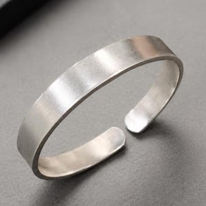 Puede incluir: Una pulsera tipo brazalete de metal color plata con un acabado mate y liso. La pulsera presenta un diseño sencillo y abierto con bordes redondeados, creando una banda continua que se curva en forma circular, mostrando una superficie uniforme.