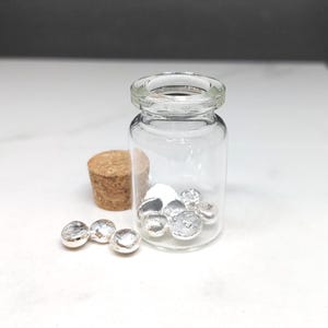 May include: A clear glass vial containing small, shiny, silver-coloured metal pieces. A cork stopper and additional metal pieces are next to the vial. The items are on a white surface.