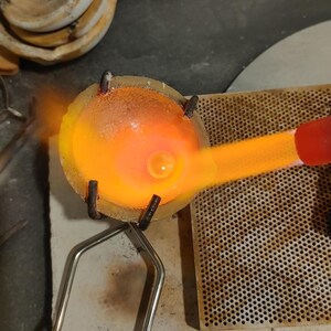 A jewelery-making setup with a crucible containing molten metal, glowing orange from the heat of a torch. The crucible is held by a metal stand, and other tools are visible in the background.