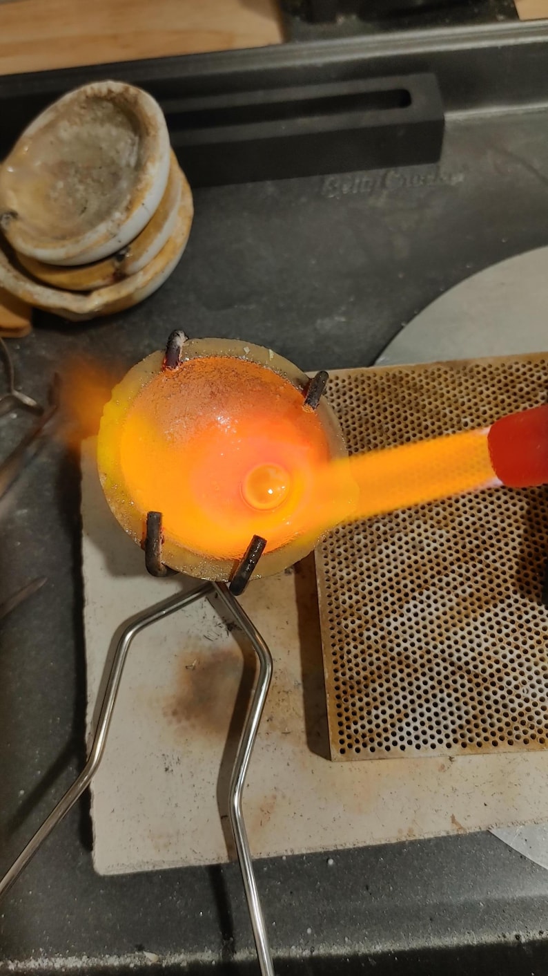 A jewelery-making setup with a crucible containing molten metal, glowing orange from the heat of a torch. The crucible is held by a metal stand, and other tools are visible in the background.
