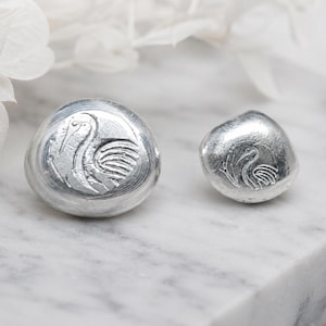 Close‑up of 999 fine silver grain shot pieces displayed on a light neutral surface beside a bamboo block. Each grain shows smooth rounded edges and bright metallic luster, highlighting purity and craftsmanship. Ideal for jewelry making, casting, or refining, these fine silver granules reflect light beautifully and emphasize the clean, modern aesthetic of artisan materials.