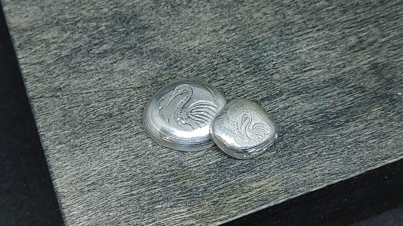 Close‑up of 999 fine silver grain shot pieces displayed on a light neutral surface beside a bamboo block. Each grain shows smooth rounded edges and bright metallic luster, highlighting purity and craftsmanship. Ideal for jewelry making, casting, or refining, these fine silver granules reflect light beautifully and emphasize the clean, modern aesthetic of artisan materials.