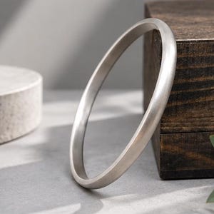 May include: A solid, circular bangle bracelet with a smooth, matte silver-toned finish. The bracelet features a simple, continuous band design with a rounded profile, reflecting light evenly across its metallic surface.