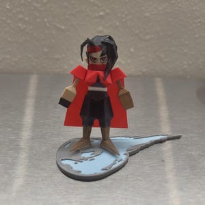 May include: A red and black 3D printed figurine of a character with a red cape and a blue sword. The figurine is standing on a blue and gray base.