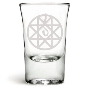 May include: A clear glass shot glass with a white etched design of a circle with a star inside and a flame on top.