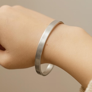 以下が含まれることがあります： シルバーカラーの平バンドブレスレット。ブレスレットはつや消しメタル仕上げで、手首に着用されています。背景はニュートラルなベージュ色です。ブレスレットはシンプルでミニマリストなデザインです。