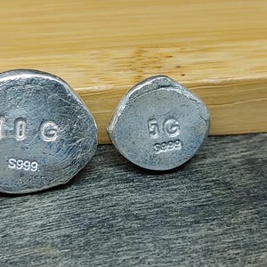 Close‑up of 999 fine silver grain shot pieces displayed on a light neutral surface beside a bamboo block. Each grain shows smooth rounded edges and bright metallic luster, highlighting purity and craftsmanship. Ideal for jewelry making, casting, or refining, these fine silver granules reflect light beautifully and emphasize the clean, modern aesthetic of artisan materials.