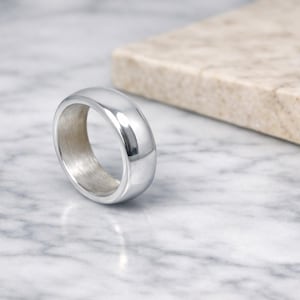 Peut inclure: Une bague argentée polie avec un anneau arrondi. La bague est posée sur une surface en marbre avec un objet rectangulaire clair en arrière-plan. La surface lisse de la bague reflète la lumière.
