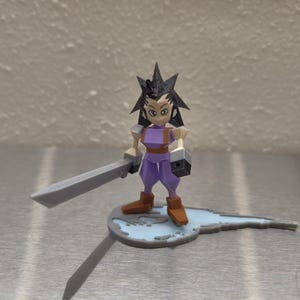 May include: A purple and gray 3D printed figurine of a character holding a sword. The character has black hair and is standing on a blue and gray base.