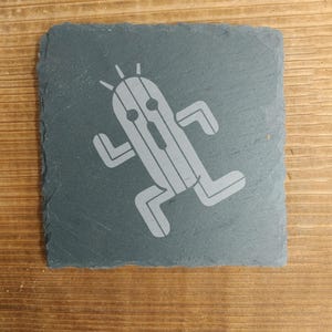 以下が含まれることがあります： 驚いた表情をしたサボテンのような生き物が走っている、ビデオゲームのキャラクターの白いシルエットが描かれた灰色のスレートコースターです。