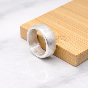 May include: A silver ring with a brushed finish rests on a light brown wooden surface. The ring has a wide band and a rounded shape. The background is a white marble surface. The ring is a piece of jewelry.