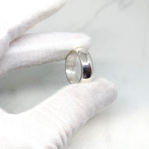 May include: A polished silver ring with a rounded band. The ring is held by a gloved hand against a white background. The ring's surface reflects light, creating highlights and shadows.