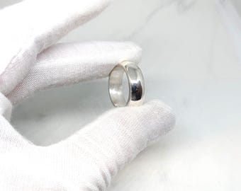 May include: A polished silver ring with a rounded band. The ring is held by a gloved hand against a white background. The ring's surface reflects light, creating highlights and shadows.