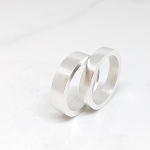 Puede incluir: Dos anillos de plata con un acabado de metal cepillado plano. Los anillos están uno al lado del otro, con uno ligeramente superpuesto al otro. Los anillos están sobre un fondo blanco.