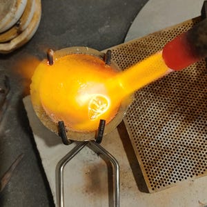 A jewelery-making scene showing a crucible with molten metal being heated by a torch. The crucible is held by metal tongs. The bright orange flame is directed at the metal. The scene is set on a perforated metal surface.