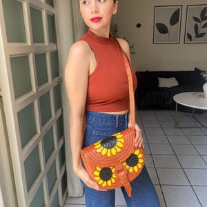 Handcrafted Mexican leather crossbody bag modeled by a woman in a rust sleeveless top and denim jeans. The bag is tan brown with hand-tooled sunflower details in bright yellow and black, featuring a curved flap, rustic stitching, and an adjustable leather strap. A unique bohemian accessory perfect for adding handmade floral charm and artisan craftsmanship to everyday outfits or travel looks.