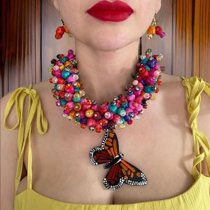 Op de afbeelding: Een kleurrijke kralenketting met een grote vlinderhanger. De ketting is gemaakt van kleine, kleurrijke kralen en heeft een grote, zwarte vlinderhanger met rode, oranje en gele accenten.