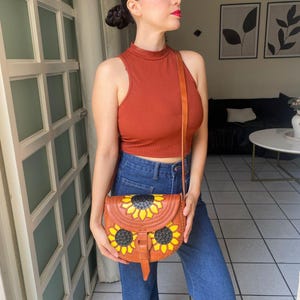 Handcrafted Mexican leather crossbody bag modeled by a woman in a rust sleeveless top and denim jeans. The bag is tan brown with hand-tooled sunflower details in bright yellow and black, featuring a curved flap, rustic stitching, and an adjustable leather strap. A unique bohemian accessory perfect for adding handmade floral charm and artisan craftsmanship to everyday outfits or travel looks.