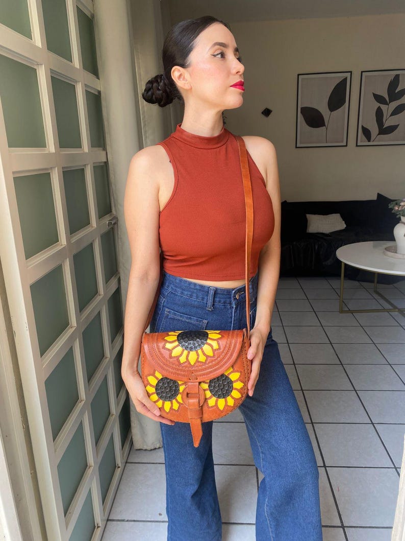 Handcrafted Mexican leather crossbody bag modeled by a woman in a rust sleeveless top and denim jeans. The bag is tan brown with hand-tooled sunflower details in bright yellow and black, featuring a curved flap, rustic stitching, and an adjustable leather strap. A unique bohemian accessory perfect for adding handmade floral charm and artisan craftsmanship to everyday outfits or travel looks.