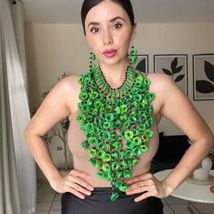 以下が含まれることがあります： 鮮やかなグリーンのフローラルネックレスとイヤリング。ネックレスは、黒いビーズのアクセントが付いたグリーンの花のクラスターが特徴です。イヤリングはフローラルデザインを反映しています。黒いスカートとヌードカラーのトップスと合わせて着用されています。