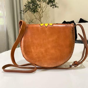 Handcrafted Mexican leather crossbody bag modeled by a woman in a rust sleeveless top and denim jeans. The bag is tan brown with hand-tooled sunflower details in bright yellow and black, featuring a curved flap, rustic stitching, and an adjustable leather strap. A unique bohemian accessory perfect for adding handmade floral charm and artisan craftsmanship to everyday outfits or travel looks.