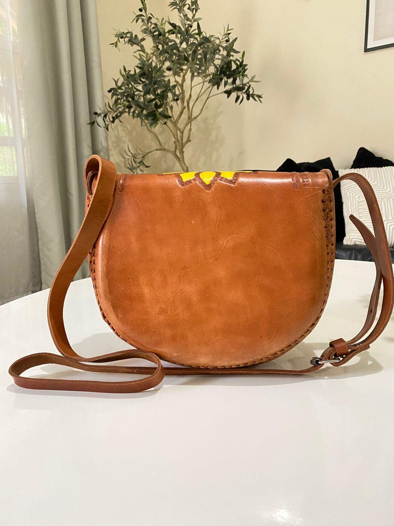 Handcrafted Mexican leather crossbody bag modeled by a woman in a rust sleeveless top and denim jeans. The bag is tan brown with hand-tooled sunflower details in bright yellow and black, featuring a curved flap, rustic stitching, and an adjustable leather strap. A unique bohemian accessory perfect for adding handmade floral charm and artisan craftsmanship to everyday outfits or travel looks.