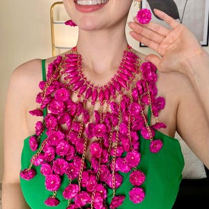 May include: A vibrant fuchsia floral necklace and matching earrings. The necklace features a cascade of fuchsia flowers and gold-colored chain accents. The woman in the image is wearing a green dress. The jewelry is a bold statement piece.