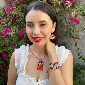 Può includere: Una donna indossa una camicetta bianca con una collana e un braccialetto di perline rosse con figure ricamate colorate. La collana e il braccialetto presentano un design tradizionale dell'arte popolare messicana.