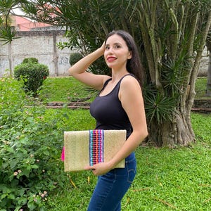 Handmade palm clutch featuring a rectangular woven palm leaf body, decorated with vibrant multicolor tassels and hand-painted pine cone flowers in pink, yellow, turquoise, purple, orange, and red. The center strip has a colorful embroidered textile accent, creating a unique artisan boho design. Perfect as a statement accessory for festivals, summer parties, vacations, or as a one-of-a-kind handmade gift for women.